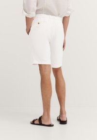 White, knee-length shorts made of cotton with an elastic waistband and a button detail. Paired with simple brown sandals on bare feet.