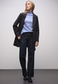 Ciemne, szerokie dżinsy z pomarańczowym przeszyciem, jasnoniebieski golf i czarny płaszcz. Model stoi na szarej podłodze na neutralnym tle.