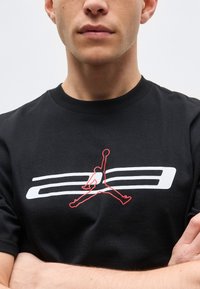 Άνδρας που φοράει μαύρο μπλουζάκι με κόκκινο και άσπρο λογότυπο Jumpman, με σταυρωμένα χέρια, σε απλό φωτεινό φόντο.