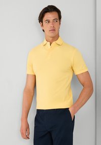 Polo tričko s krátkým rukávem ve světle žluté barvě, se klasickým límcem a knoflíkovou légou na tři knoflíky, v kombinaci s tmavě modrými kalhotami.