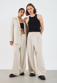 Veste de costume beige ajustée, crop top noir et pantalons beige à jambes larges. Chaussures noires texturées. Deux mannequins se tenant côte à côte.