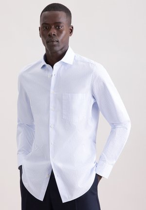 Mand iført lyseblå og hvide tyndstribede skjorte med knapper, lange ærmer og mørke bukser, stående mod en ensartet lys baggrund.