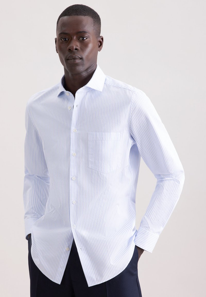 Homme portant une chemise à fines rayures bleu clair et blanc, à boutons et manches longues, ainsi qu'un pantalon foncé, debout devant un fond clair uni.