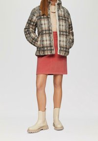 Veste en flanelle à carreaux duveteuse en crème, beige et noir ; col roulé beige clair ; jupe en velours côtelé rose ; bottines beiges à semelles épaisses et texturées.
