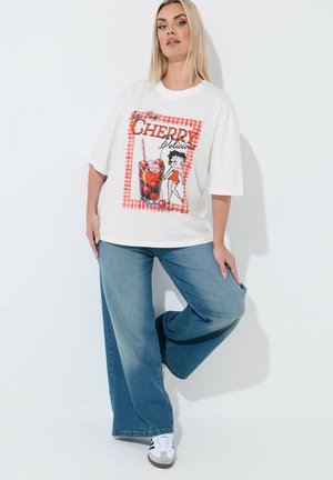 Donna con capelli biondi che indossa una t-shirt bianca oversize con una grafica di una bevanda alla ciliegia e un personaggio dei cartoni animati, jeans blu a gamba larga e sneakers bianche.