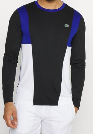Homme portant une chemise à manches longues avec un panneau central noir, des accents bleus aux épaules, des côtés blancs et un petit logo de crocodile vert sur la poitrine.