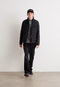 Zwarte pufferjas met een rits, gedragen over een grijs shirt, gecombineerd met donkere broek en zwarte schoenen. Een zwarte pet met het woord "BOSS."