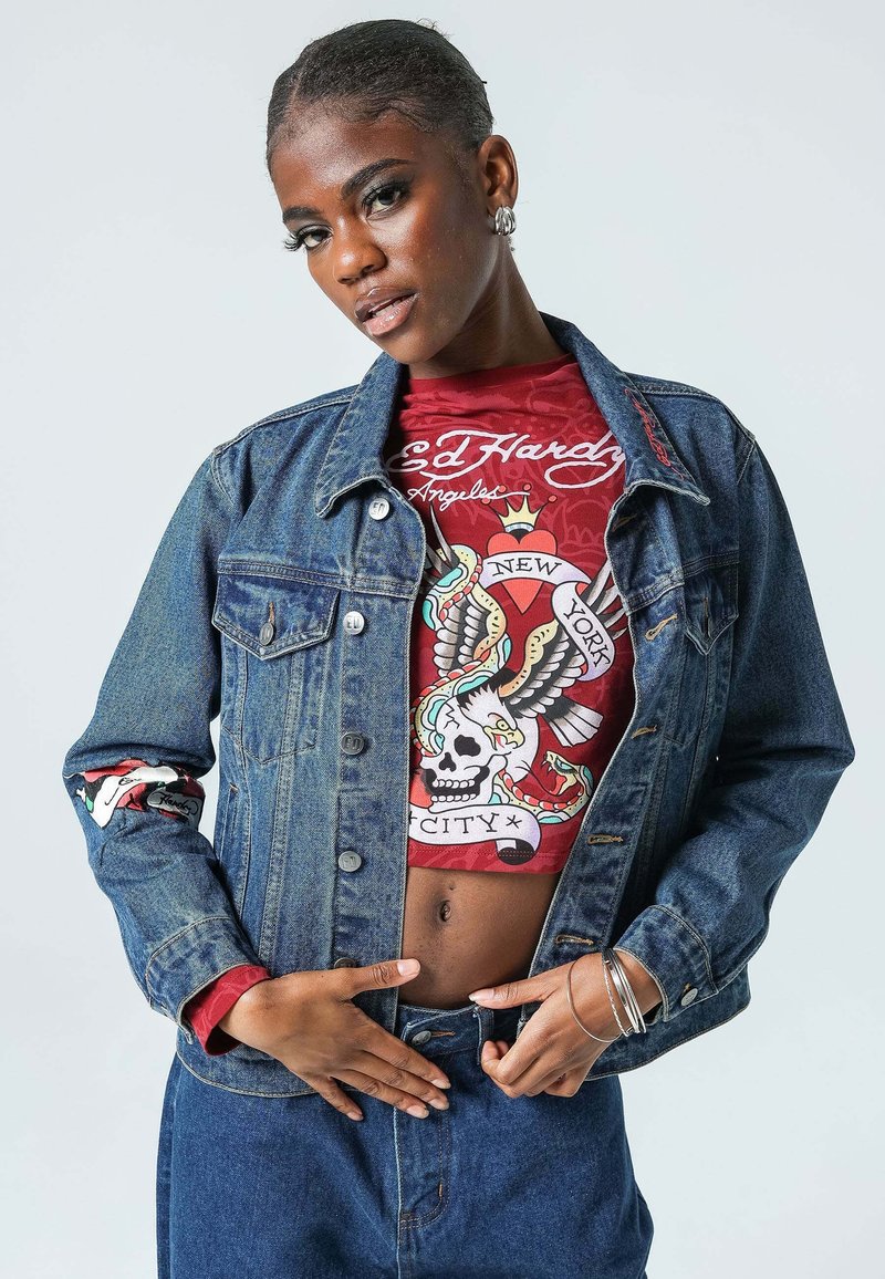 Chaqueta de mezclilla con botones en el frente, dos bolsillos en el pecho y un acento rojo en las mangas, superpuesta sobre una camiseta gráfica roja con diseños llamativos.