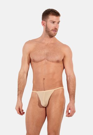 Beiger Nylon-String mit dünnen Trägern; glatte Textur; minimale Bedeckung; getragen von einem Mann, stehend posierend, mit kurzen Haaren und Bart.