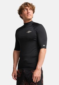 Rashguard noir à manches courtes avec un col montant, arborant un petit logo sur la poitrine. Le matériau semble lisse avec une coupe ajustée.