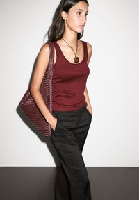 Débardeur bordeaux en coton, pantalon noir ajusté avec poches, et un sac tote tissé bordeaux. Collier pendentif avec une pierre marron.
