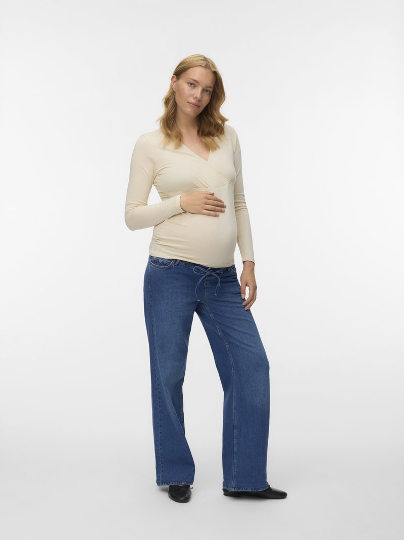 Beige wikkelshirt met lange mouwen over een zwangere buik, gecombineerd met losse blauwe spijkerbroek en zwarte platte schoenen; gladde textuur en casual ontwerp.