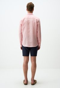 Leichtes, strukturiertes Langarmhemd in Pink mit dezenten Streifen, kombiniert mit dunklen Shorts und Sandalen. Die Rückansicht zeigt den Kragen und die Länge des Shirts.