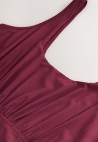 Sleeveless burgundy top with gathered fabric detail below the shoulder strap on a light background.