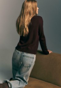 Kobieta z długimi blond włosami, ubrana w ciemny sweter i luźne jasnoniebieskie dżinsy, opierająca się na oliwkowo-zielonej kanapie, patrzy sideways.
