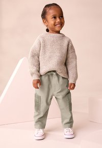 Maglione beige a coste, pantaloni cargo verde chiaro con tasche in velluto a coste e sneaker bianche. Bambino in piedi su una superficie morbida.