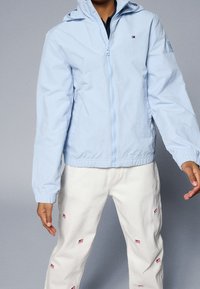 Světle modrá bunda na zip s kapucí, elastickými manžetami a lemem. S detaily loga, doplněná bílými kalhotami s malými motivy.