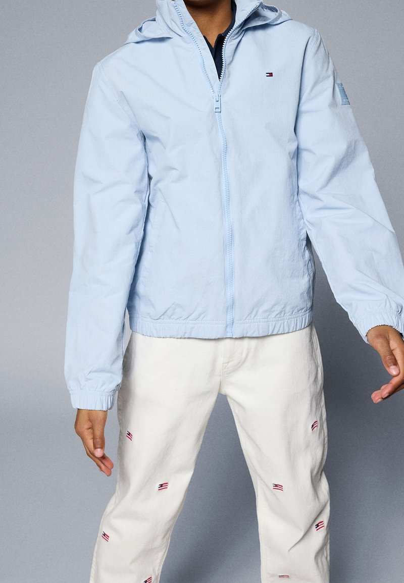 Světle modrá bunda na zip s kapucí, elastickými manžetami a lemem. S detaily loga, doplněná bílými kalhotami s malými motivy.