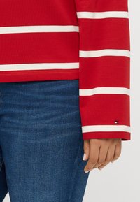 Haut rouge à manches longues avec des rayures horizontales blanches, fabriqué en tissu doux. Présente un petit logo sur la manche et un col rond.