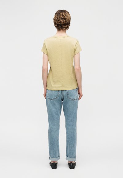 Άτομο με κοντά σγουρά μαλλιά που φοράει ανοιχτόκίτρινο πουκάμισο με κοντό μανίκι, ανοιχτό μπλε τζιν με γυρισμένα μπατζάκια και μαύρα παπούτσια, στέκεται γυρισμένο πλάτη.