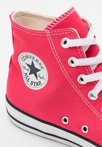 Sapatilha de lona rosa de cano alto com atacadores brancos, apresentando um patch de logotipo de estrela em preto e branco e riscas pretas na sola de borracha.