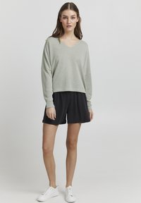 Lichtgroene, losvallende gebreide trui met een V-hals, gecombineerd met zwarte, zachte shorts. Witte sneakers maken de ontspannen outfit compleet.