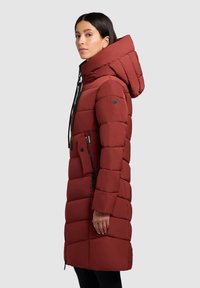 Mujer vestida con un abrigo acolchado largo color óxido con gran capucha y bolsillos con cremallera, de pie en perfil frente a un fondo liso.
