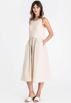Femme portant une robe midi beige sans manches avec taille froncée et poches latérales, associée à des sandales blanches à talons ouverts, debout sur un fond blanc.