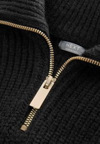 Pull noir texturé zippé avec une fermeture éclair dorée et une étiquette indiquant "NEXT, FABRIQUÉ EN CHINE." Vue rapprochée mettant en avant le matériau et les éléments métalliques.