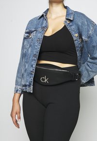 Černá ledvinka Calvin Klein se stříbrným logem, elegantní design a zipové zapínání. Nositá přes texturované černé legíny a džínovou bundu.