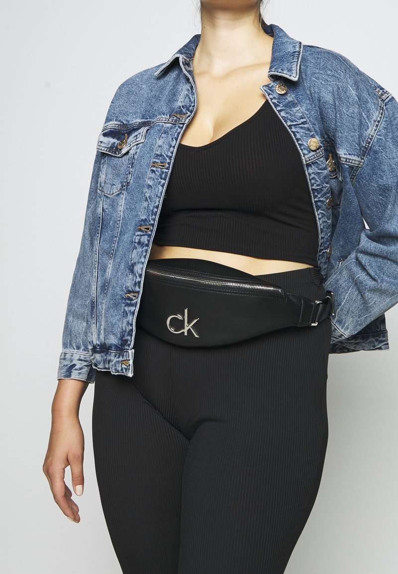 Černá ledvinka Calvin Klein se stříbrným logem, elegantní design a zipové zapínání. Nositá přes texturované černé legíny a džínovou bundu.