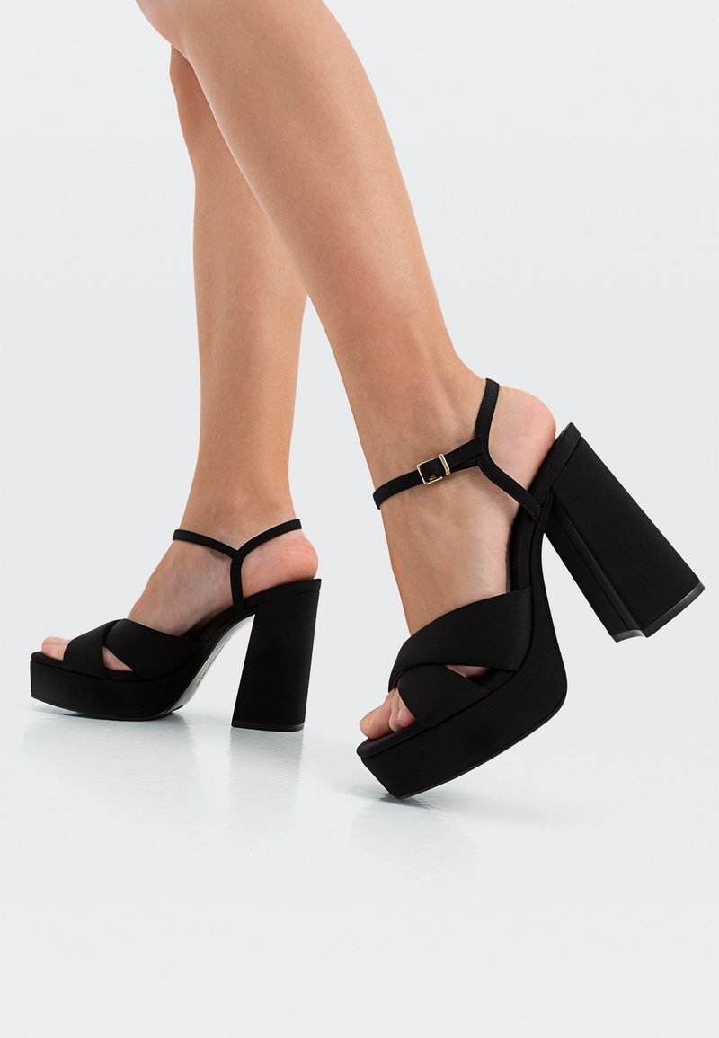 Sandales à plateforme noires avec un design de bride texturé présentant un motif entrecroisé et un talon épais. Fermeture à boucle à la cheville.