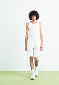Hvid tanktop og lysegrå shorts med en lyserød logo-detalje. Model iført hvide sneakers, stående på et grønt tæppe foran en ensfarvet baggrund.