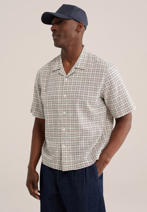 Homme portant une casquette de baseball bleu marine, une chemise à manches courtes boutonnée avec un motif à carreaux rouge et bleu, et un pantalon bleu foncé, debout avec une main dans la poche.