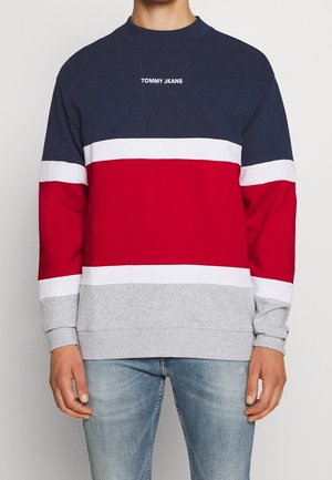 Mężczyzna w bluzie z blokami kolorystycznymi w granatowe, białe, czerwone i szare paski oraz logo „TOMMY JEANS” na piersi, w połączeniu z jasnoniebieskimi jeansami.