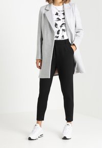 Cappotto lungo grigio sopra una t-shirt grafica bianca, pantaloni neri svasati e sneakers bianche. Tessuto morbido, design su misura e accenti minimalisti.