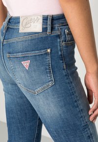 Jeans de mezclilla azul con un diseño ajustado, que presentan un lavado claro, bolsillos traseros y un parche de logo con strass en la cinturilla.