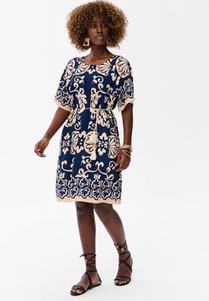 Kobieta w granatowej sukience do kolan z beżowymi kwiatowymi wzorami, paskiem z frędzlami, dużymi kolczykami, bransoletkami i czarnymi sandałami na sznurowadła.