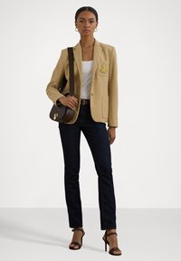 Blazer beige avec un patch, haut blanc, jeans en denim foncé et sac à main marron. Des sandales à talons complètent la tenue. Tissu lisse et coupe ajustée.