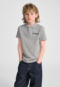 Szara koszulka polo z kołnierzykiem i trzema guzikami, z czarnym logo na lewej piersi, zestawiona z niebieskimi dżinsami.