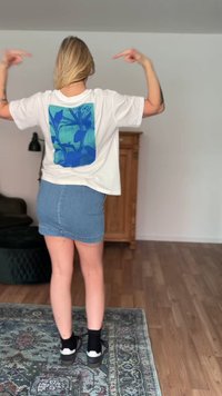 Γυναίκα στέκεται μέσα σε κλειστό χώρο και δείχνει το σχέδιο μπλε φυτού στην πίσω πλευρά της λευκής μπλούζας της, φοράει τζιν φούστα και μαύρα αθλητικά παπούτσια.