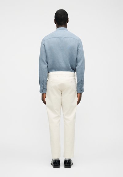 Homme debout, dos tourné, portant une chemise à manches longues bleu clair, un pantalon crème, des chaussettes grises et des chaussures habillées noires, sur un fond blanc uni.