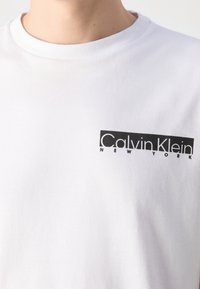 Gros plan d'une personne portant un t-shirt blanc à col rond avec un logo noir rectangulaire Calvin Klein New York sur la poitrine gauche.
