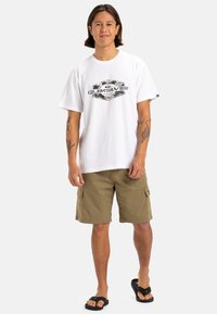 Camiseta blanca de algodón con el logo negro de Quiksilver en el pecho, combinada con pantalones cortos de carga beige con bolsillos laterales y chanclas negras.
