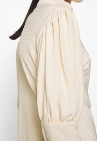 Kermavärinen pitkähiha blouse, jossa on tekstuuri, rakenteelliset olkapäät ja keräilyt yksityiskohdat hihoissa.