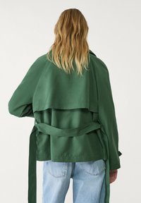 Grøn, langærmet jakke i glat stof med lagdelt ryg, bindebælte i taljen og afslappet pasform. Bæres over lyseblå jeans.