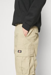 Pantaloni cargo beige con finitura testurizzata, dotati di tasche laterali, vita regolabile e un'etichetta di marca sulla coscia.