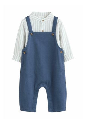 Salopetă din denim albastru cu bretele reglabile, nasturi din lemn și o decupare rotundă în partea de jos. Sub aceasta este un tricou cu mânecă lungă, în dungi albe.