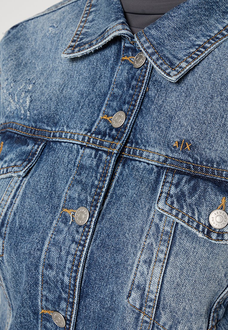 Chaqueta de mezclilla en azul claro, con un cuello clásico, cierres de botón plateados, acentos de costura en naranja y dos bolsillos en el pecho.