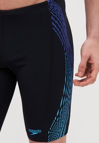 Sorte svømmebukser laget av elastisk materiale, med et blått og turkist bølgete mønster langs siden, synlige sømmer og logo.
